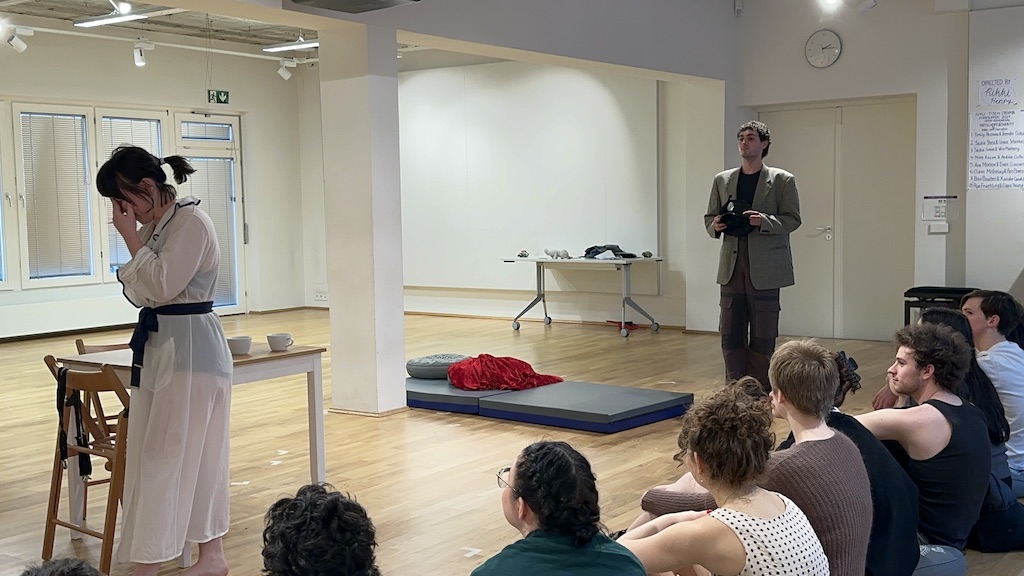 Two actors stand facing each other in mid-performance of 'Faith Hope Charity'. A table and chairs are set in front of the actors with a mattress and red blanket to their right.